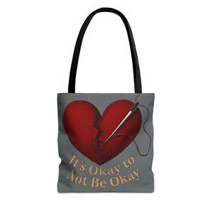 May include: Gray tote bag with a black handle featuring a red heart graphic with a visible crack being sewn together with a needle and thread. The text "It's Okay to Not Be Okay" is printed in gold.