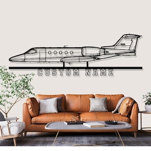 May include: Black metal airplane wall art with the text "CUSTOM NAME" below the plane. The airplane is a detailed outline of a jet. The art is mounted above a brown leather sofa with pillows.