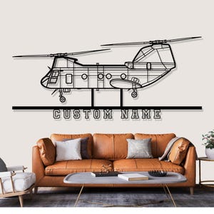 May include: Black metal helicopter wall art with the text "CUSTOM NAME" below the aircraft. The helicopter features detailed outlines of the body, rotors, and windows. The artwork is displayed above a brown leather sofa.