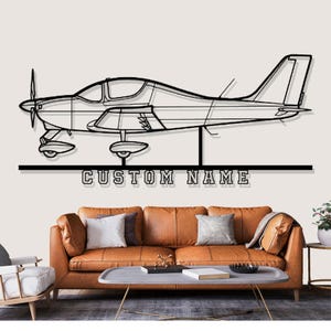 Op de afbeelding: Zwarte metalen vliegtuig wandkunst met de tekst "CUSTOM NAME" onder het vliegtuig. Het vliegtuig heeft gedetailleerde contouren van de cockpit, vleugels en propeller. De kunst wordt tentoongesteld boven een bruine leren bank.