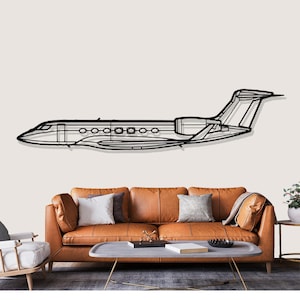 Puede incluir: Arte mural de metal negro de un jet privado. La obra es un contorno del perfil lateral de la aeronave, con ventanas, motores y cola detallados. Montado en una pared de color neutro sobre un sofá de cuero marrón.