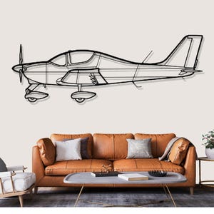 Op de afbeelding: Zwart metalen wandkunst die een klein vliegtuig afbeeldt. Het kunstwerk toont een gedetailleerde omtrek van het vliegtuig, inclusief de propeller, vleugels en staart. Het vliegtuig is gemonteerd op een neutraal gekleurde muur boven een bruine leren bank.