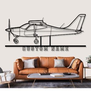 Op de afbeelding: Zwarte metalen vliegtuig wandkunst met de tekst "CUSTOM NAME" onder het vliegtuig. Het vliegtuig heeft een propeller, vleugels en een staart. De kunst wordt weergegeven boven een bruine leren bank.