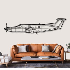 Könnte beinhalten: Schwarze Metall-Flugzeug-Wandkunst. Das Kunstwerk zeigt eine detaillierte Umrisszeichnung eines Propellerflugzeugs. Das Flugzeug ist an einer neutralfarbenen Wand über einem braunen Ledersofa mit Kissen und einem Couchtisch angebracht.