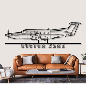 Könnte beinhalten: Schwarze Metall-Wandkunst mit einer Flugzeugsilhouette mit Propeller und dem Text "CUSTOM NAME" darunter. Das Kunstwerk ist über einem braunen Ledersofa mit Kissen und einem Couchtisch angebracht.