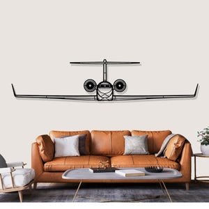 Puede incluir: Una pieza de arte mural de avión de metal negro. La obra de arte representa una silueta detallada de un avión a reacción, con alas, cola y detalles del motor. El arte está montado sobre un sofá de cuero marrón.