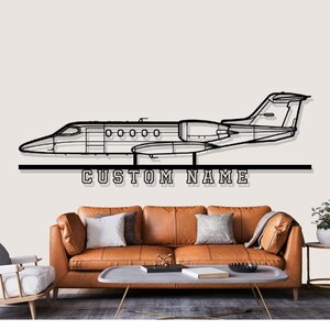 May include: Black metal wall art depicting a detailed outline of a private jet. The text "CUSTOM NAME" is below the jet. The art is mounted above a brown leather sofa with decorative pillows.