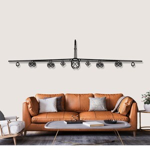 May include: A black metal wall art piece of an airplane, mounted above a brown leather sofa. The artwork shows the silhouette of the aircraft, including wings, engines, and fuselage. The room has a neutral colour palette.