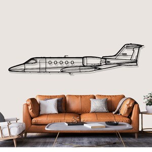 May include: A black and white line drawing of a private jet is mounted on a wall above a brown leather sofa. The jet has five windows and a detailed outline of the plane's body and wings.