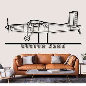 May include: Black metal airplane wall art with the text "CUSTOM NAME" below the plane. The airplane is a side profile with a propeller and detailed wings. The art is displayed above a brown leather sofa.