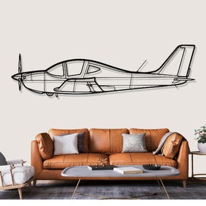 Op de afbeelding: Zwarte metalen vliegtuig wandkunst. Het kunstwerk toont de omtrek van een klein vliegtuig met een propeller, vleugels en staart. Het vliegtuig is gemonteerd op een neutraal gekleurde muur boven een bruine leren bank.