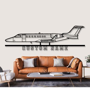 May include: Black metal wall art depicting an airplane silhouette with the text "CUSTOM NAME" below. The artwork is mounted above a brown leather sofa with pillows and a coffee table.
