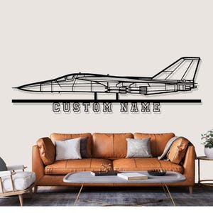 Peut inclure: Décoration murale en métal noir représentant un avion à réaction avec le texte "CUSTOM NAME" en dessous. L'œuvre est au-dessus d'un canapé en cuir marron avec des coussins et une table basse.