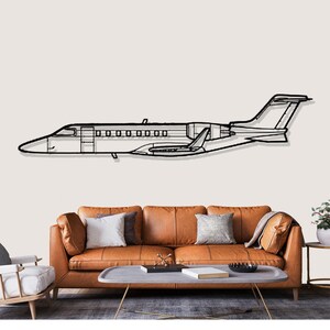 May include: Black metal wall art of a private jet. The outline of the aircraft is visible, including the fuselage, wings, and tail. The artwork is mounted on a light-colored wall above a brown leather sofa.