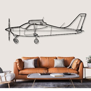 Op de afbeelding: Zwarte metalen vliegtuig wandkunst. Het vliegtuig is een zijaanzicht met een propeller, vleugels en staart. Het kunstwerk is gemonteerd op een lichtgekleurde muur boven een bruine leren bank met kussens.