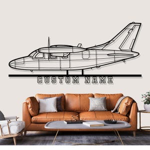 May include: Black metal airplane wall art with the text "CUSTOM NAME" below the plane. The airplane is a detailed outline of a small aircraft. The art is displayed above a brown leather sofa.