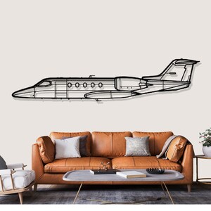 May include: Black metal wall art depicting a jet airplane. The artwork is a line drawing of the plane's silhouette, with details of the windows, wings, and tail. The art is mounted on a light-colored wall above a brown leather sofa.