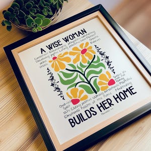 May include: Framed art print with the text "A Wise Woman Builds Her Home." The print features a floral design with yellow and red flowers, green leaves, and black accents. The frame is black and the matting is beige.