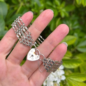 Puede incluir: Una pulsera plateada con un cierre de candado en forma de corazón. La pulsera presenta un diseño tejido y una pequeña cadena que conecta el candado. El fondo es una vista borrosa de follaje verde y flores blancas.