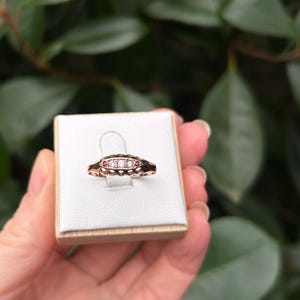 May include: A rose gold ring with three clear stones in the center and two red stones on the sides. The ring is displayed on a white surface, held by a hand, with a green leafy background.