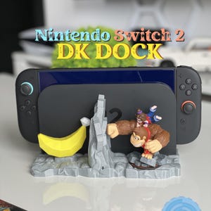 Può includere: Un dock Nintendo Switch 2 DK a tema Donkey Kong. Il dock è grigio con una banana gialla e una statuina di Donkey Kong. Il testo "Nintendo Switch 2 DK DOCK" è visualizzato in alto.