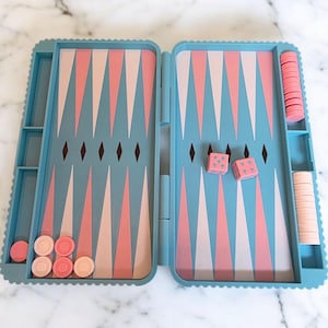 Könnte beinhalten: Ein Backgammon-Set im Vintage-Stil in einem hellblauen Koffer mit einem rosa-weißen Dreiecksmuster auf der Spielfläche. Enthält rosa Würfel und Spielsteine. Das Set ist geöffnet und spielbereit.