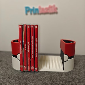 May include: A white and red video game cartridge holder with a ridged base, holding several red game cases. The holder has a design reminiscent of a popular animated series. The text "Printaættis" is visible in the background.