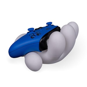 Könnte beinhalten: Ein blau-schwarzer Videospiel-Controller liegt in einem weißen, handförmigen Halter. Der Controller hat blaue Knöpfe und einen schwarzen Mittelteil. Der Halter ist glatt und abgerundet, mit modernem Design.