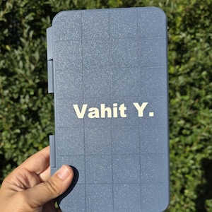 Könnte beinhalten: Eine blau-graue rechteckige Hülle mit einem Rastermuster und dem Text "Vahit Y." in Weiß. Die Hülle hat einen Klappverschluss und wird in einer Hand vor einem Hintergrund aus grünem Laub gehalten.