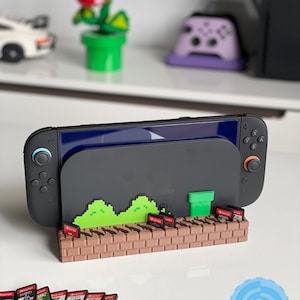 Può includere: Una console Nintendo Switch nera con un supporto personalizzato con un design a mattoni pixelati e paesaggio verde. Il supporto contiene cartucce di gioco. Una statuina di pianta Piranha e altri accessori sono visibili.