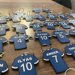 Peut inclure: Plusieurs porte-clés en forme de maillot bleu avec des bordures blanches, chacun portant un nom et un numéro. Les porte-clés sont disposés sur une surface marron, avec des noms comme "AMIN" et des numéros comme "6".