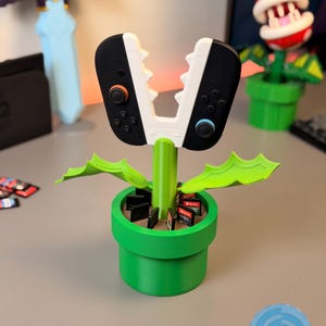 Può includere: Un vaso verde stampato in 3D a forma di tubo, che contiene un controller per videogiochi e cartucce di gioco. Il controller è bianco e nero, simile alla bocca aperta di una pianta. Il vaso ha foglie verdi e uno stelo verde.