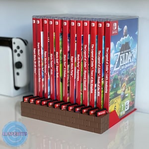 Op de afbeelding: Een bruine spelcartridgehouder in baksteenstijl met meerdere rode spelhoezen. De hoezen zijn gelabeld met speltitels als "The Legend of Zelda" en "Super Mario". Een witte spelconsole is zichtbaar op de achtergrond.