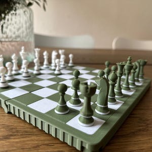 Op de afbeelding: Een groen en wit schaakspel met alle stukken op hun plaats. Het schaakbord is groen met witte vierkanten. De schaakstukken zijn wit en groen. De set staat op een houten oppervlak.