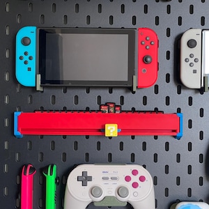 Puede incluir: Un panel perforado negro con varios accesorios de juego. Una consola Nintendo Switch con mandos azules y rojos está montada, junto con un soporte rojo para cartuchos de juego. Otros artículos incluyen un mando blanco y mandos Joy-Con rosas y verdes.