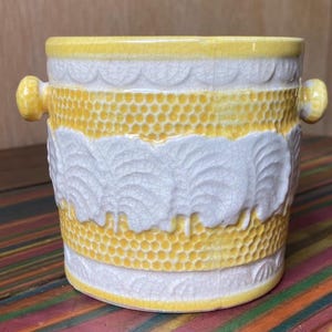May include: A ceramic planter with a yellow and white design. The planter has a yellow rim and handles, with a textured pattern of yellow dots and white leaf-like shapes. The planter is approximately 5 inches tall.