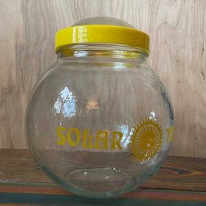 May include: A clear glass jar with a yellow lid and rim. The jar has the word "SOLAR" in yellow lettering with a smiling sun graphic. The jar is sitting on a wooden surface.
