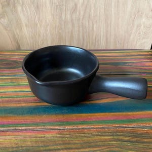 May include: A small, black ceramic saucepan with a single handle. The pan has a rounded shape and a matte finish. The handle is curved and extends from the side of the pan. The background is a wooden surface.