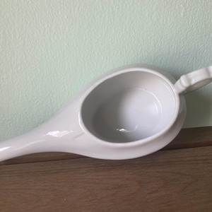 May include: A white ceramic neti pot, featuring a rounded bowl, a spout, and a small handle. This nasal irrigation device is shown on a wooden surface.
