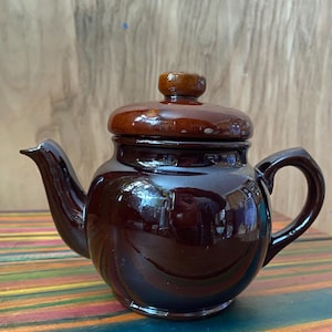 May include: A brown ceramic teapot with a matching lid and handle. The teapot has a glossy finish and a spout for pouring. The lid has a small, round knob. The teapot is sitting on a colorful striped surface.