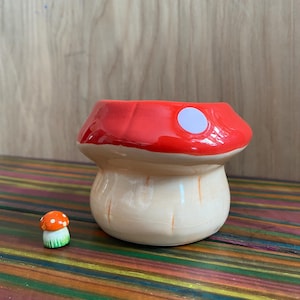 May include: A ceramic mushroom-shaped planter with a red cap and a white dot. The stem is off-white with vertical lines. A miniature mushroom figurine with an orange cap and white dots sits nearby. The planter is a whimsical decorative item.