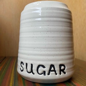 Può includere: Un contenitore per lo zucchero in ceramica bianca con finitura maculata e texture a coste orizzontali. La parola "SUGAR" è in rilievo in nero sulla parte anteriore del contenitore. Il contenitore ha una forma cilindrica.