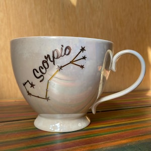 May include: A pearlescent white ceramic mug with a gold-colored "Scorpio" text and constellation design. The mug has a rounded shape, a handle, and a small pedestal base. The background is a wooden surface.
