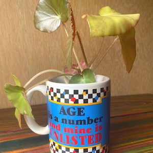 May include: A white ceramic mug featuring a black and white checkered border with small colorful geometric shapes. The blue center panel displays the text "AGE is a number and mine is UNLISTED" in blue and red. A green plant grows from the mug.