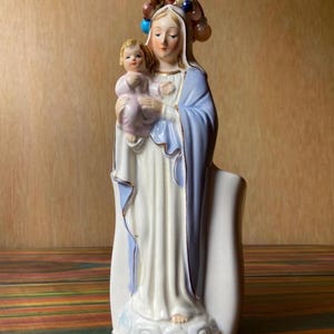 May include: A ceramic figurine of the Virgin Mary holding a child. The statue is predominantly white with blue and gold accents. The Virgin Mary wears a crown of beads and holds the child in a flowing robe. The base is decorated with gold trim.