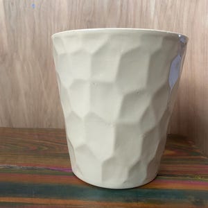 May include: A cream-colored ceramic planter with a geometric, textured surface. The cylindrical pot has a wide opening and sits on a multi-colored wooden surface. Ideal for indoor plants.