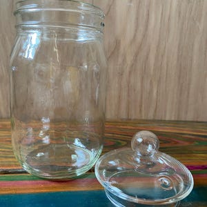 May include: A clear glass jar with a matching lid. The jar is cylindrical with a wide mouth and a screw-on rim. The lid is oval-shaped with a small, round knob on top. The jar and lid are made of transparent glass.