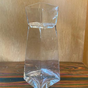 Peut inclure: Un vase en verre transparent de forme géométrique avec un design angulaire unique. Le vase a une base large en forme de losange qui se rétrécit vers le haut jusqu'à une ouverture carrée. Le verre transparent laisse passer la lumière.