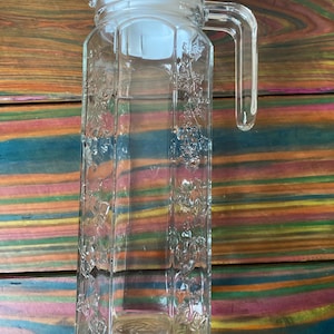 Puede incluir: Una jarra de vidrio transparente con tapa y asa de plástico blanco. La jarra tiene forma rectangular con diseños florales en relieve. El asa es transparente y curvada. La jarra está vacía y sobre una superficie de madera.