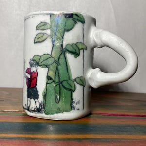 May include: White ceramic mug with a curved handle. The mug features a hand-drawn illustration of a person playing golf next to a green bamboo stalk with leaves. The mug is sitting on a wooden surface.
